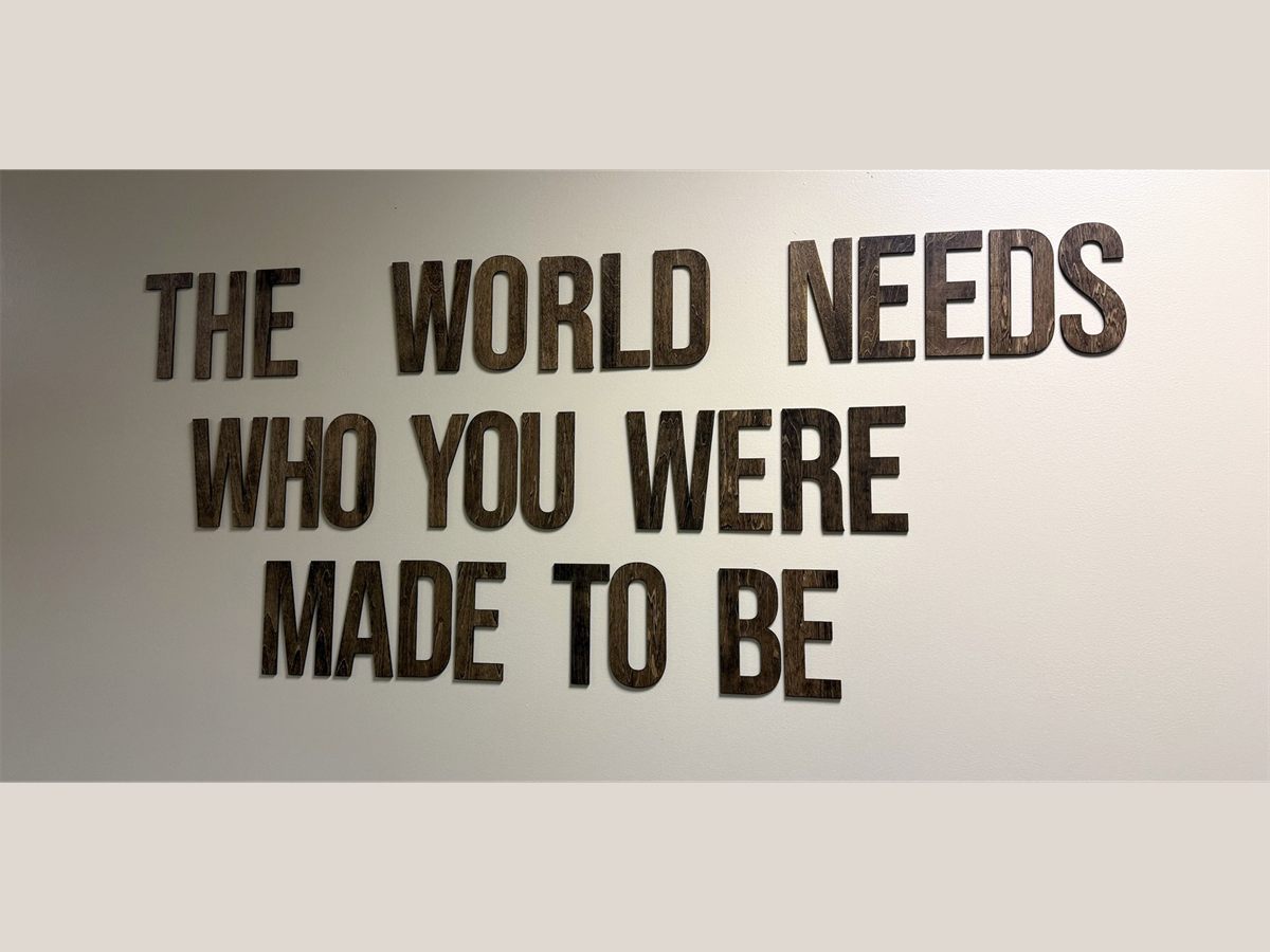 Stained plywood letters reading 'The World Needs Who You Were Made To Be' mounted on a wall