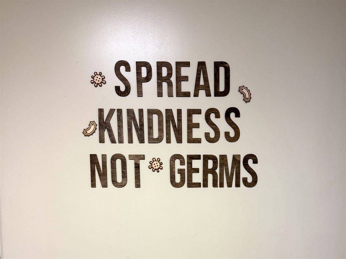 Stained plywood letters reading 'Spread Kindness Not Germs' on a white wall, with engraved germ-shaped accents